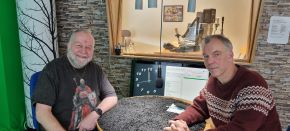 Två män sitter vid ett bord i en radiostudio. Till vänster sitter Gunnar Cronenberg med skägg, iklädd en mörk t-shirt med ett tryck. Till höger sitter Stefan Clint i en rödbrun mönstrad stickad tröja. Bakom dem syns studioutrustning, en datorskärm och en grön skärm samt en stenmönstrad vägg.